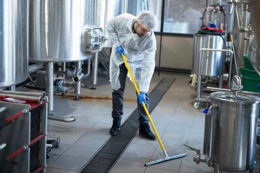 Industrial cleaner in protective coveralls mopping the floor in a factory with steel tanks